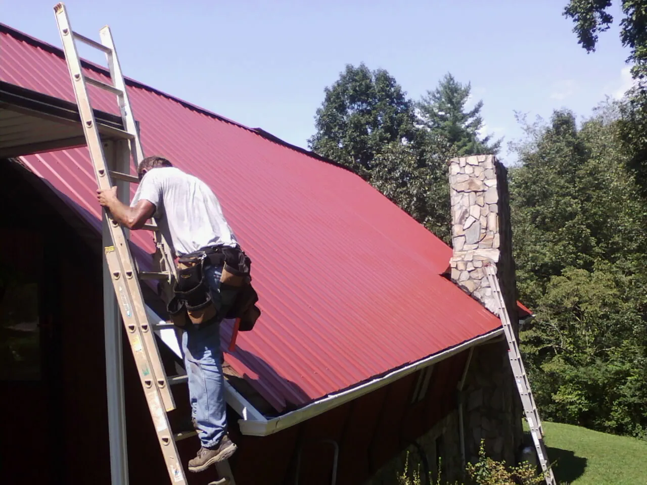 Roofer on a ladder beside a red metal roof and stone chimney — Soffit & Fascia Repair by Roofing Brothers in New Woodstock